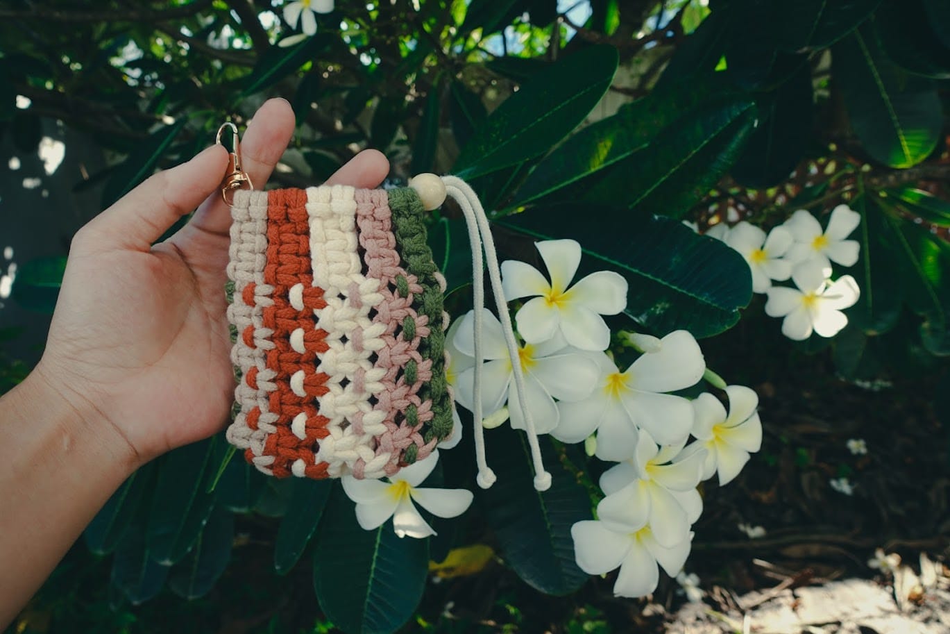 Handmade macrame pouch in ivory, terracotta, and green cords with boho style, displayed outdoors with white plumeria flowers – Macrame Pouch Video Tutorial